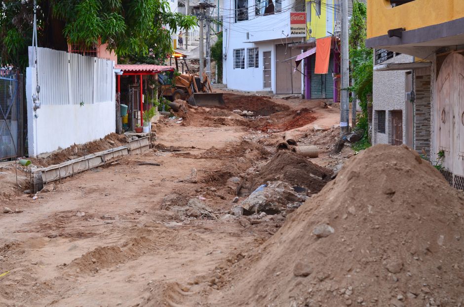 The image shows a city street undergoing roadwork, with a section of the road excavated and covered with black soil and debris. Bright orange plastic barriers and metal fencing delineate the construct